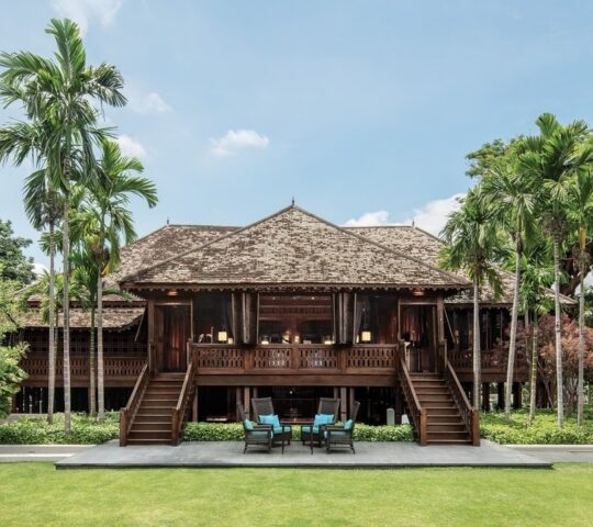 A large, traditional dark wood Thai building with twin staircases and a symmetrical facade surrounded by palm trees.