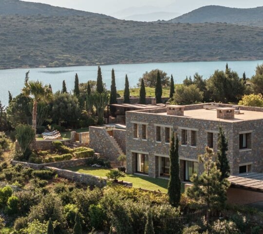 Aerial view of a villa at Phaea Blue Palace in Crete with surrounding trees and water in the background