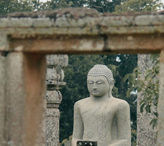 A grey stone statue of a seated Buddha viewed through a rectangular stone doorway of an ancient ruin.