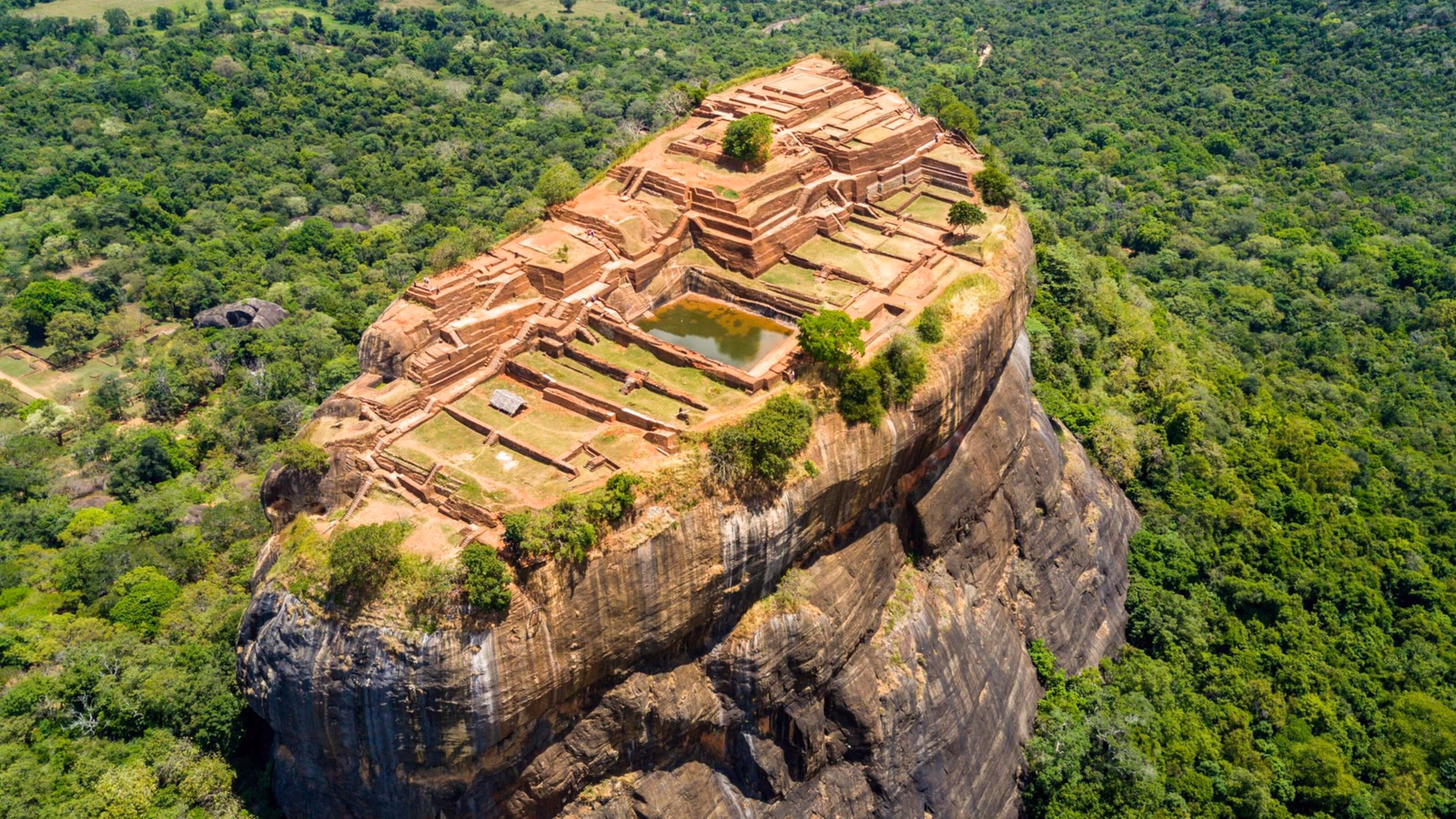 Top-down view of the flat summit of a massive rock showing foundation ruins and a small green pool.