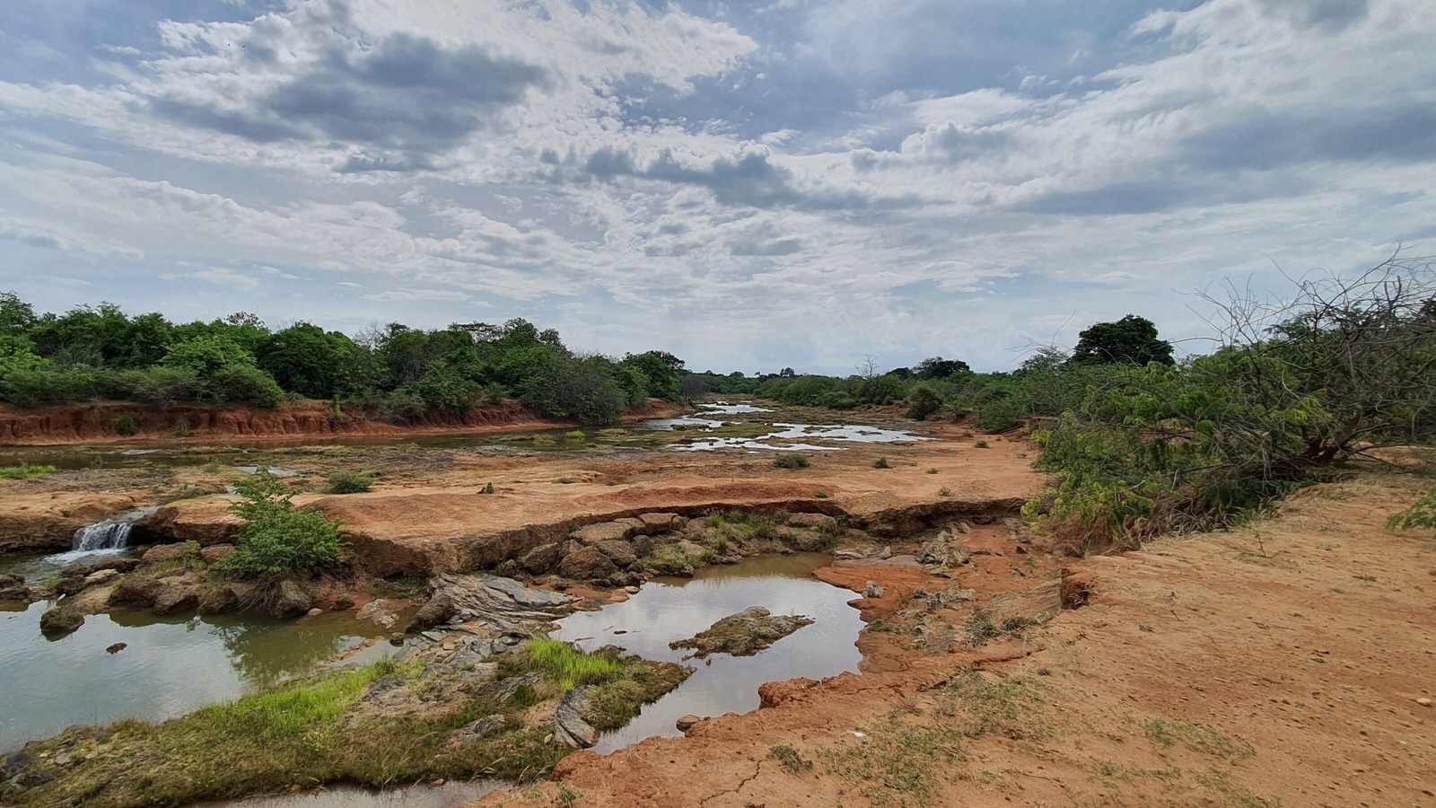 A rocky riverbed with small pools of water surrounded by red soil and green bushes under a vast sky.