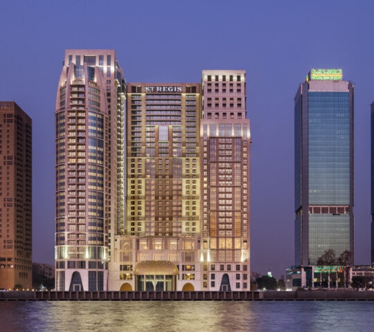 The St. Regis Cairo hotel and neighboring skyscrapers reflecting in the Nile River at dusk under a deep blue sky.