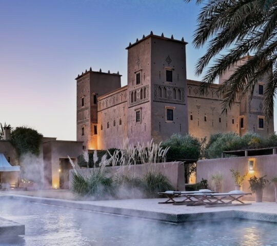 A steaming swimming pool at dusk with a large, lit-up Moroccan kasbah and palm trees in the background.