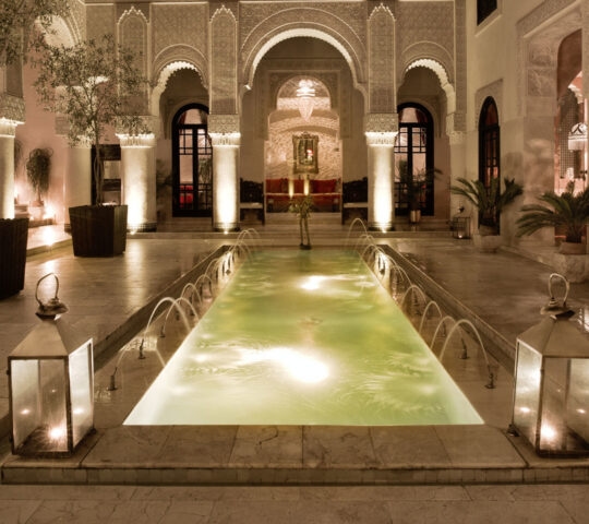 An ornate Moroccan courtyard at night with a glowing narrow pool, water fountains, and arched architecture.