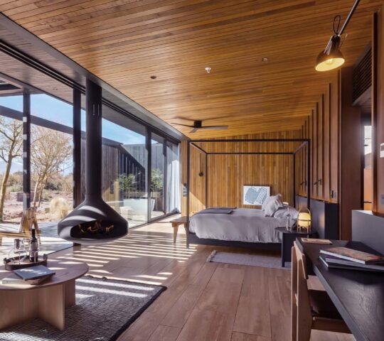 A modern luxury hotel room with wood walls, a four-poster bed, and a suspended black fireplace by large glass doors.