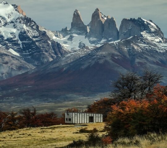 A small wooden building in a vast field with towering, snow-covered jagged mountains in the background.