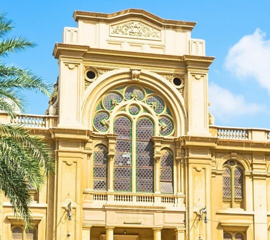 Front view of a grand sandstone synagogue with a large arched stained-glass window and surrounding palm trees.