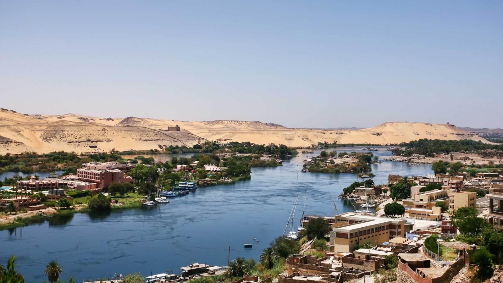 The Nile River with green islands, small boats, and sand dunes in the background under a pale sky.