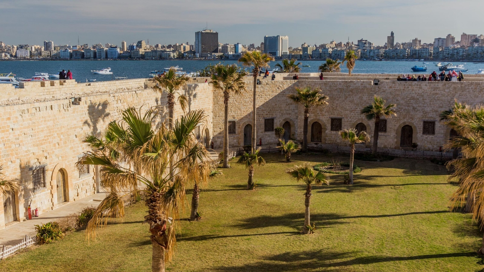 Palm trees grow in the grassy courtyard of a stone fortress overlooking the Alexandria harbor in Egypt.