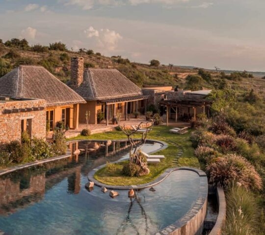 A stone luxury villa with a large, uniquely shaped infinity pool overlooking a wide, sunlit savanna landscape.