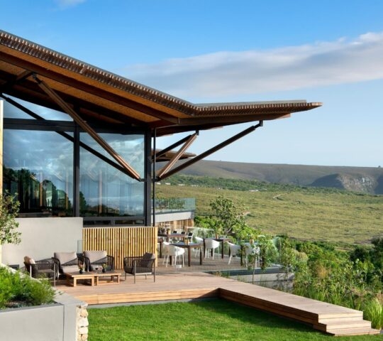 A modern glass-walled lodge with a wooden deck and outdoor seating overlooking a vast green valley under a blue sky.