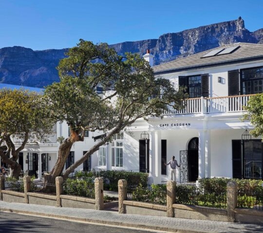 A white two-story boutique hotel with black shutters, captioned Cape Cadogan, with Table Mountain in the background.
