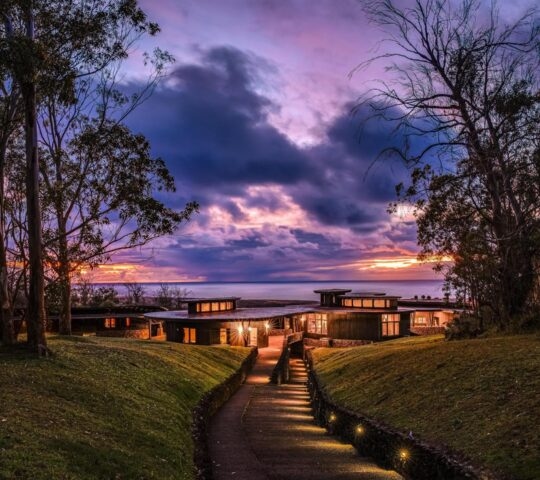 A lighted modern hotel building at dusk with a stone path leading toward it under a vibrant purple and orange sunset sky.