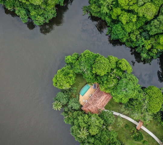 Aerial view of a luxury villa with a pool and red roof nestled in thick green jungle foliage beside a dark river.
