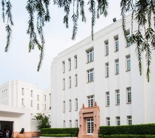The white exterior of The Imperial Delhi India with a tan stone doorway and manicured green bushes along the base.