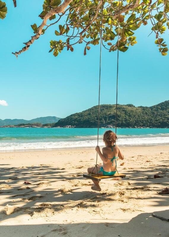 A child on a swing at a sandy beach with turquoise water and green hills in the distance in Brazil.