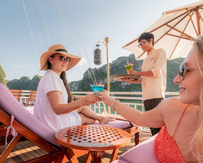 Two women toasting with blue and green cocktails on a sun deck overlooking the scenic islands of Halong Bay.