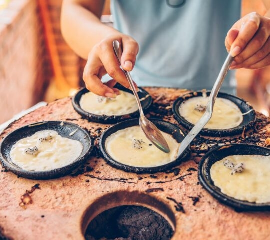 Close-up of hands using spoons to cook small rice batter pancakes in individual pans over hot coals.