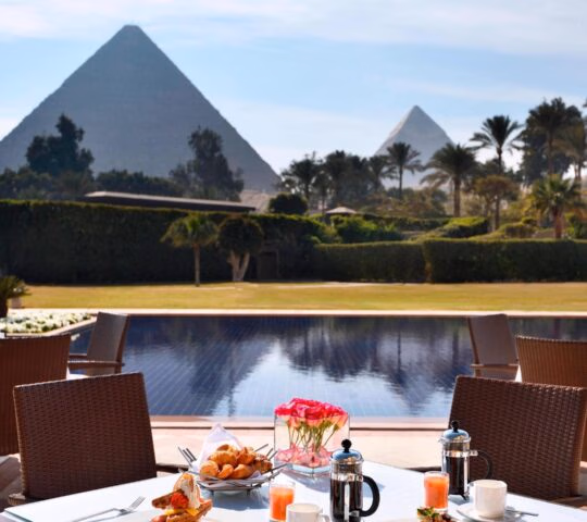 A breakfast table with pastries and coffee overlooks a swimming pool with the Giza Pyramids in the background.