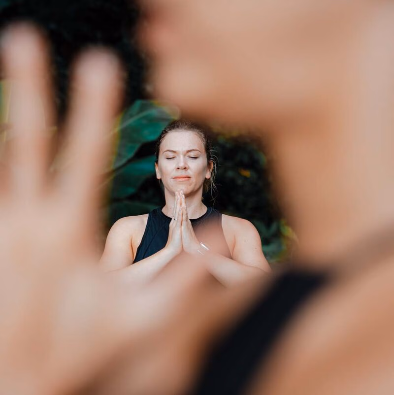 A woman closes her eyes during a yoga pose and looks peaceful with green foliage behind her and the yoga teacher in front of her.
