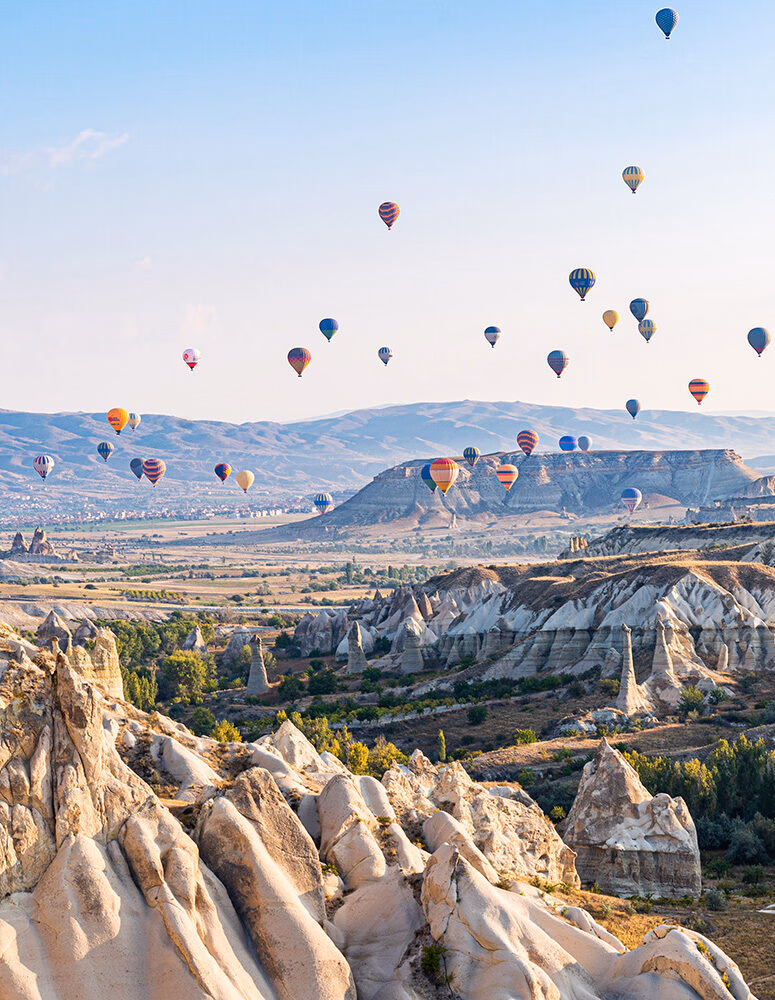 Dozens of colorful hot air balloons fly over rocky fairy chimney formations and rolling hills under a clear blue sky.