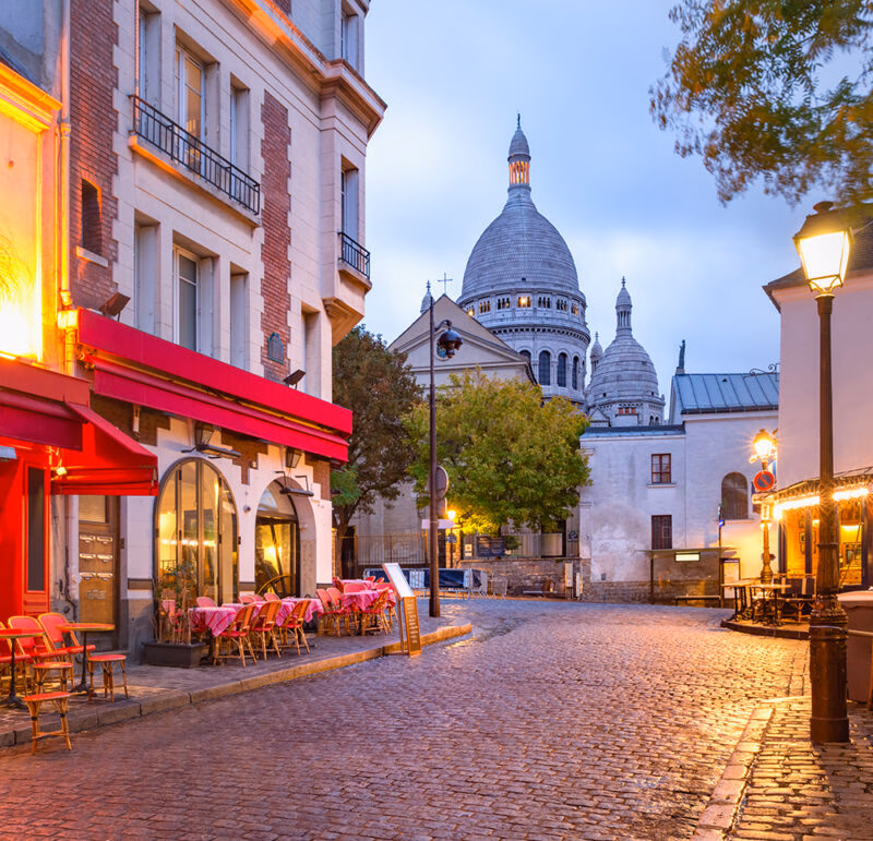 A Parisian street cafe with red chairs and tables on a cobblestone road, with a large domed basilica in the background.