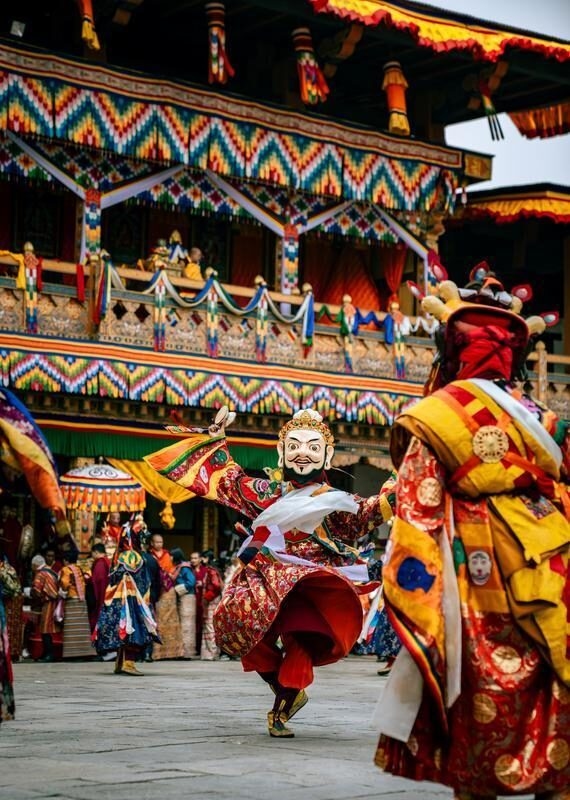 A dancer wearing a bearded mask and ornate robes performs in a courtyard.