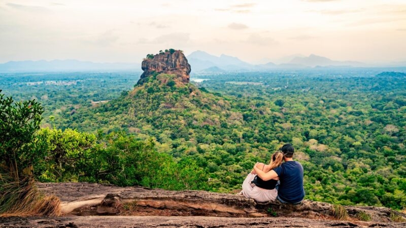 A couple sits on a ledge overlooking a vast forest toward Sigiriya rock.