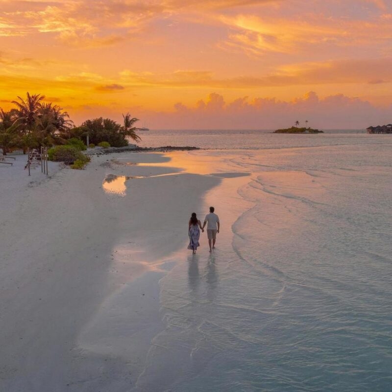 A couple walks along the edge of the ocean on a beach at sunset.