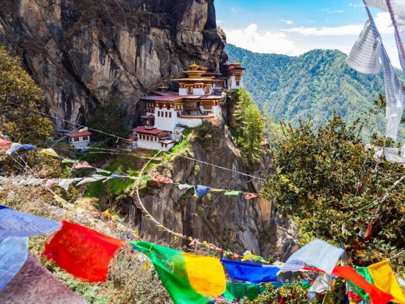 A cliffside monastery built into a steep rock face behind colorful hanging prayer flags.