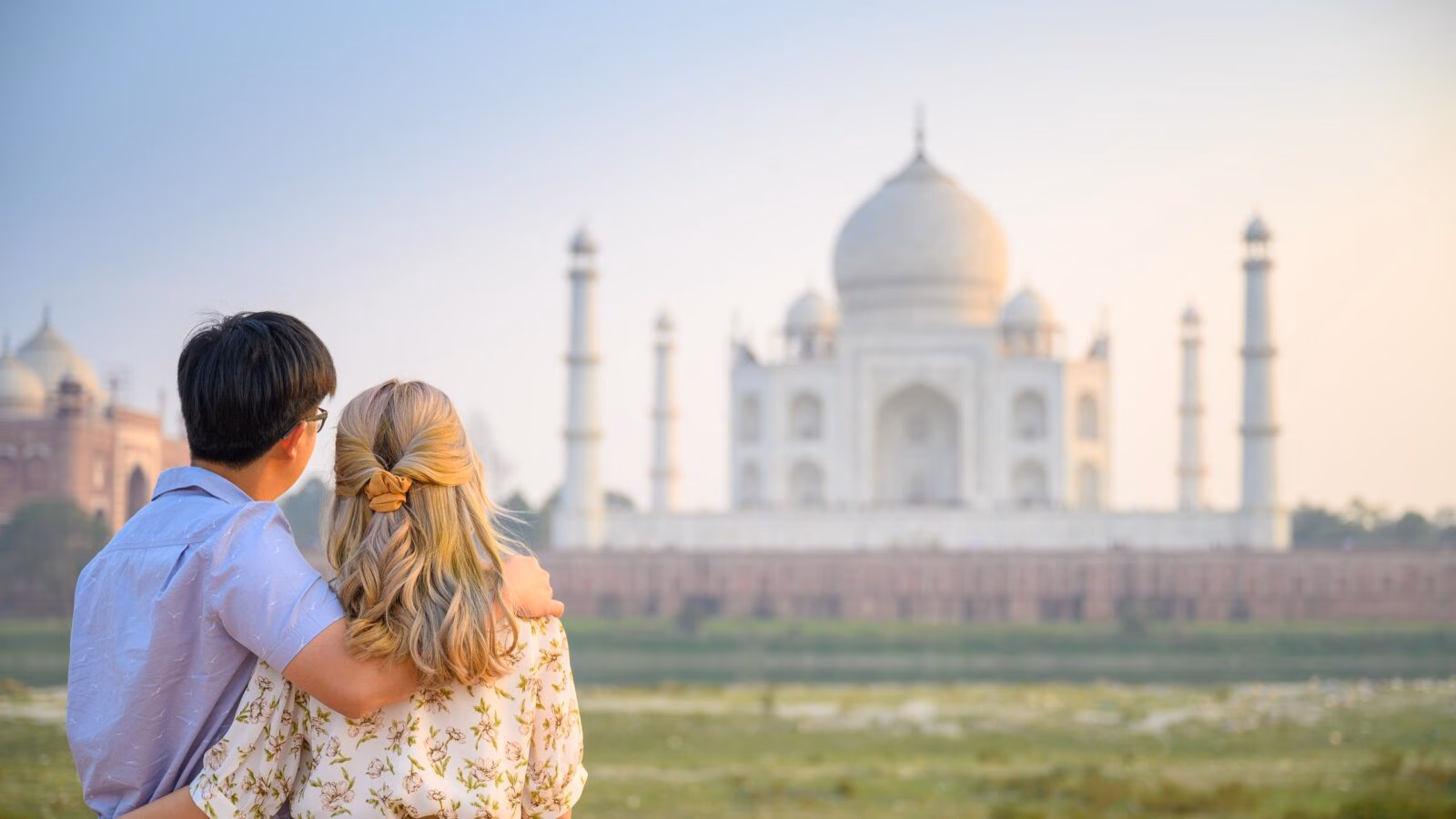 Rear view of a couple with arms around each other looking at the Taj Mahal in the distance.