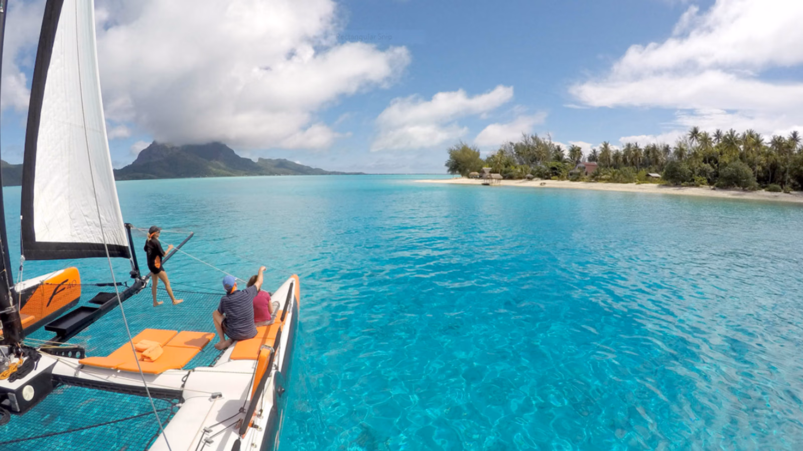 People on a catamaran sailing through clear turquoise water toward a tropical island coastline.
