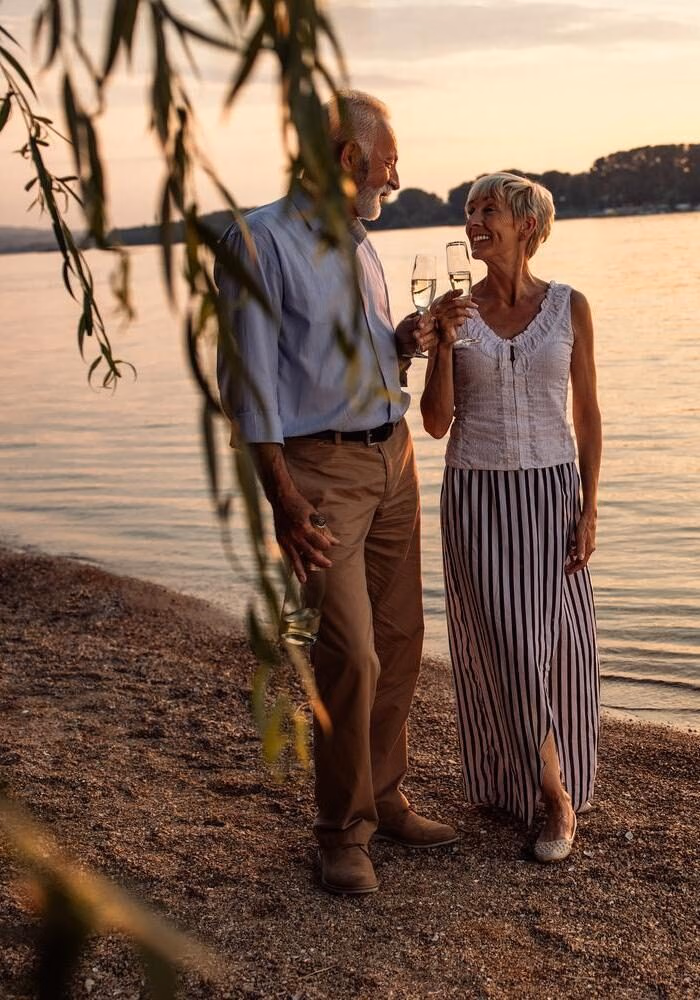 A couple holding wine glasses and smiling at each other on a beach at sunset.
