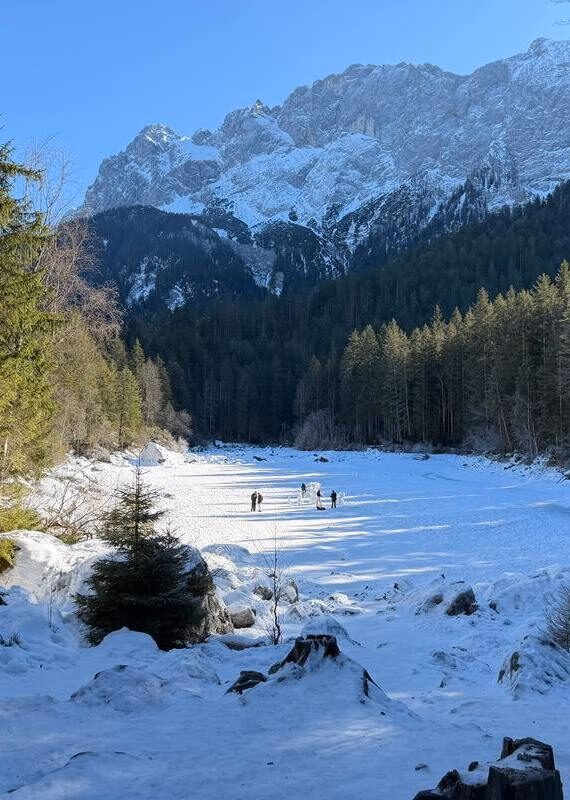 A snowy alpine valley in Bavaria with small figures walking near a pine forest under majestic winter mountains.