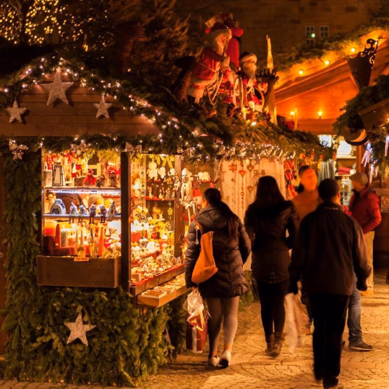 People browsing a brightly illuminated Christmas market stall filled with festive ornaments and decorations at night.