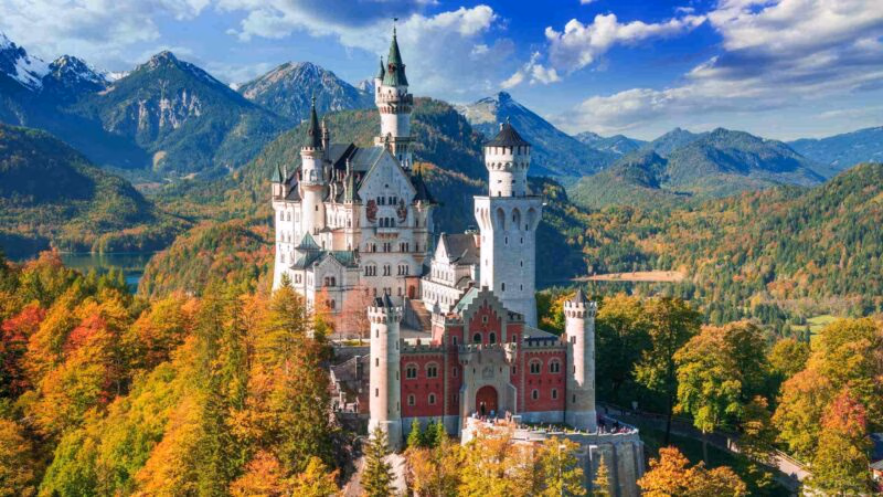 Neuschwanstein Castle surrounded by colorful autumn trees with mountains in the background.