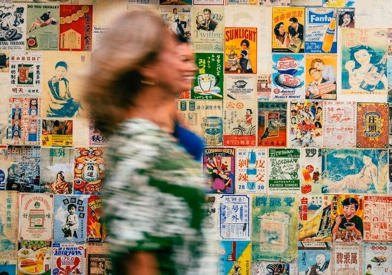 A mother and daughter on a trip exploring Bangkok's Chinatown walk past a wall adorned with vintage posters.