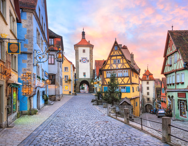 A cobblestone street splits between colorful half-timbered buildings and stone towers under a sunset sky.