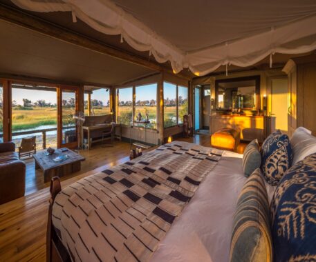 Interior image of a bedroom in a wilderness resort. There is a large bed with a canopy, a living area with a sofa and small wooden table, and view of the African landscape