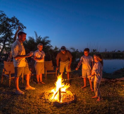 People roasting marshmallows on sticks around a campfire by an African watering hole