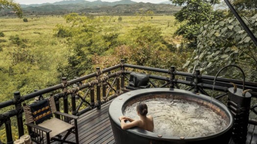 A woman sitting in a large, round hot tub on a wooden veranda looking out at a green valley with forested mountains on the horizon