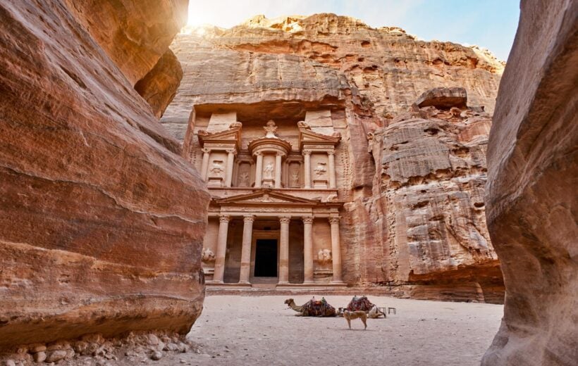 The temple-mausoleum of Al Khazneh in the ancient city of Petra in Jordan - a pinkish brown temple with columns craved out a tall craggy rock face. On the sand in front of the temple lies two camels with blankets and saddles, alongside a standing dog