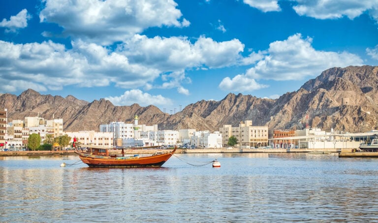 Old traditional sailboat anchored at Muttrah Corniche. The old city with white and sand coloured cube-shaped buildings and mountains are in the background. From Muscat, Oman.