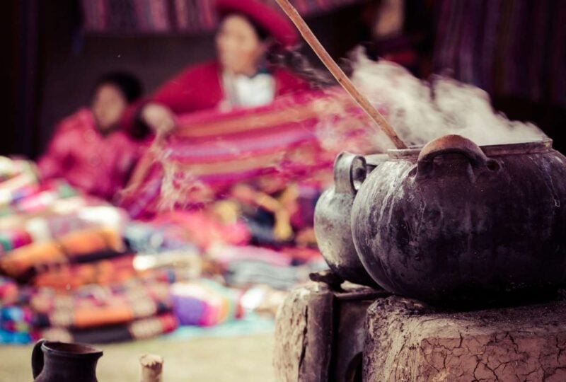 Steaming pot used in the traditional process of dyeing alpaca fur to make yarn for weaving with two Patacanchan people with their finished weaves out of focus in the background, Sacred Valley, Peru