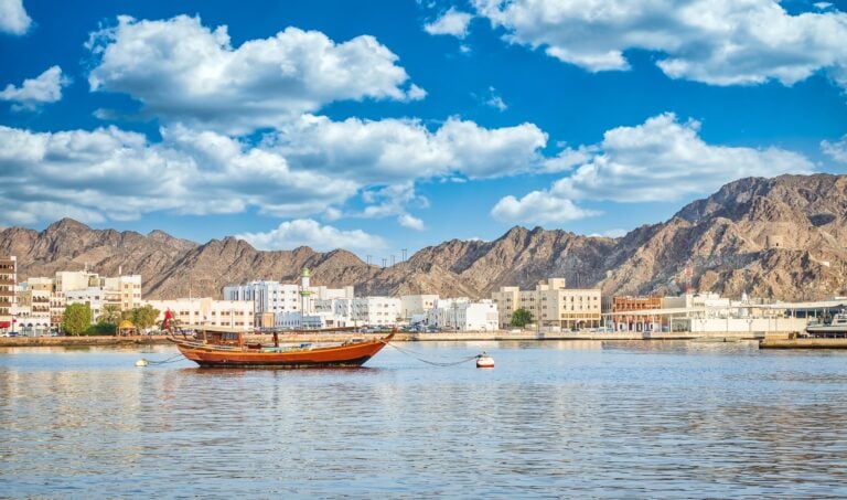 Old traditional sailboat anchored at Muttrah Corniche. The old city with white and sand coloured cube-shaped buildings and mountains are in the background. From Muscat, Oman.