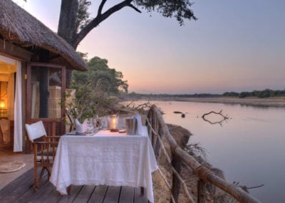 Exterior image of outdoor dining area, with small table and two chairs, set with a white table cloth, ice bucket, glasses and a candle. The view looks over a still river