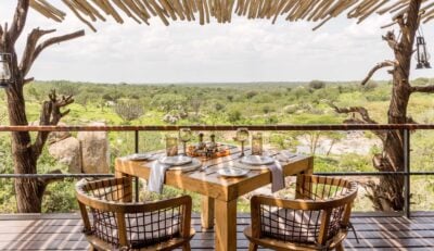 Outdoor dining area on an elevated verandah, with a square wooden table set with plates and glasses, and two chairs, overlooking the African bush