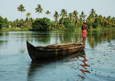 A man stands on the bow of a canoe with a long paddle steering it through palm-lined water in Kerala, India