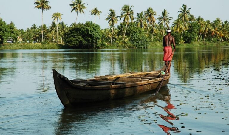 A man stands on the bow of a canoe with a long paddle steering it through palm-lined water in Kerala, India