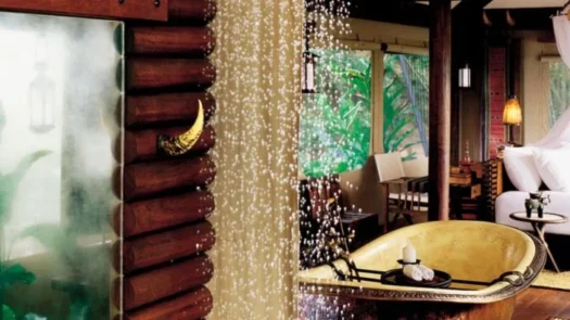 Bathroom of a luxury hotel room, with rainfall shower, a freestanding bath tub, and large bed visible in the background